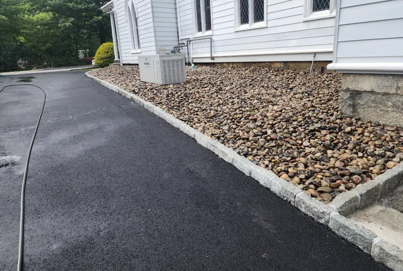A freshly laid blacktop driveway running past a white clapboard house, edged with a course of granite cobble against a bed of river stone.