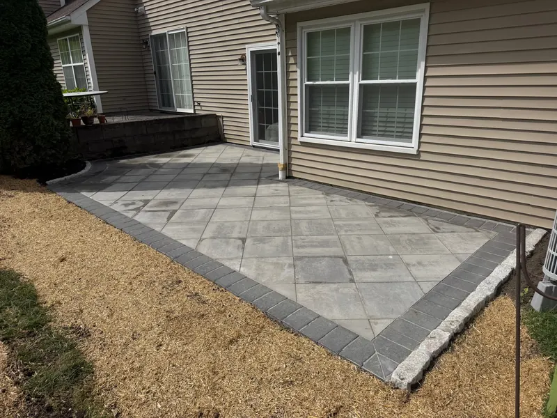 A rectangular patio in large-format grey pavers with a charcoal soldier-course border, laid against the back of a tan vinyl-sided house, with fresh straw over seeded ground along its edge.
