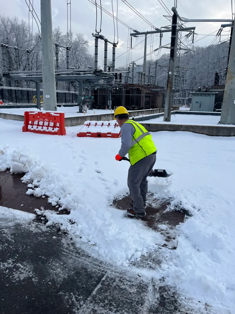 A worker in a hard hat and hi-vis clearing snow from a walkway inside a fenced electrical substation.