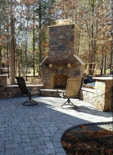 A stone outdoor fireplace with a timber mantel and an arched firebox, set behind a curved paver patio with two swivel chairs and a stone seat wall.
