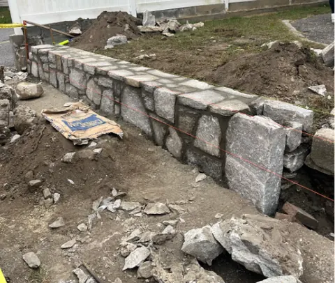 A granite block retaining wall being built to a string line, with cut stone stacked ready and the trench still open.