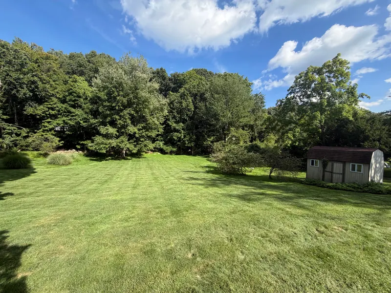A wide, freshly mown lawn with visible mower stripes running back to a mature treeline, with ornamental grasses and shrub beds along its edges and a small barn-roofed shed at the right.