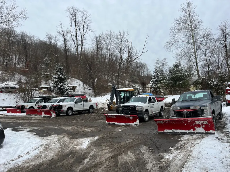 Five plow trucks with blades down, a backhoe and a spreader staged in a snow-covered yard before a storm.