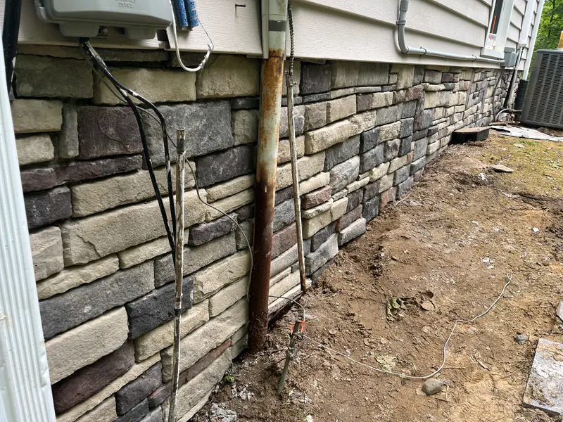 Stone veneer laid over a block foundation wall along the side of a house, with the trench still open at its base.