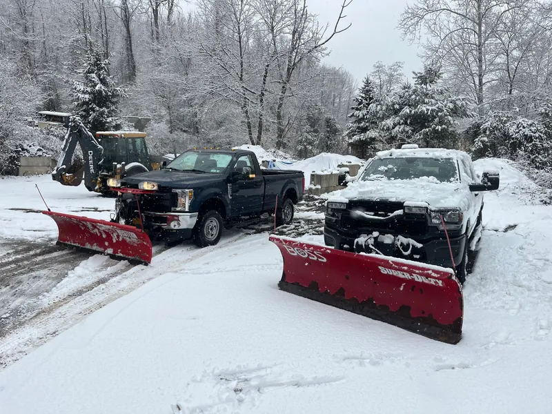 Two plow trucks with blades down on fresh snow, with a backhoe behind them and snow still falling.